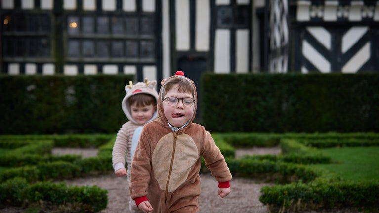 Two children dressed in reindeer onesies walk through a formal garden in front of a black and white Tudor house.